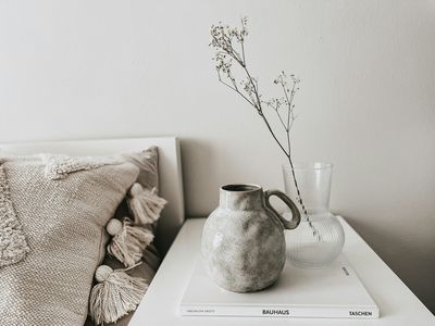 A textured stoneware pitcher and a clear glass vase with dried stems on a bedside table beside linen bedding.