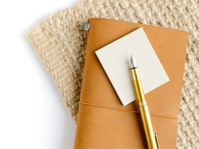 A tan leather-bound notebook and brass fountain pen resting on a woven jute mat.