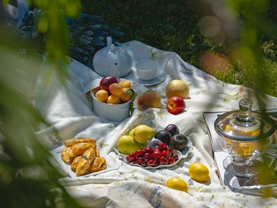A picnic laid out on a pale blanket in the grass with fruit, pastries and a glass jar, framed by green leaves.